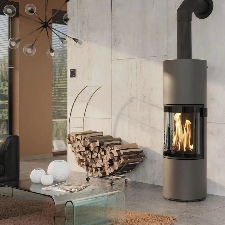 Kaminofen CERA DESIGN Prego | 6 kW – Runder Kaminofen mit Rauchrohr neben einem Holzkorb im Wohnraum