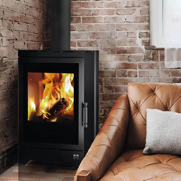 Kaminofen Hitze Lynx B 6,5 kW – Kaminofen mit Feuer neben Ledersessel und Ziegelwand