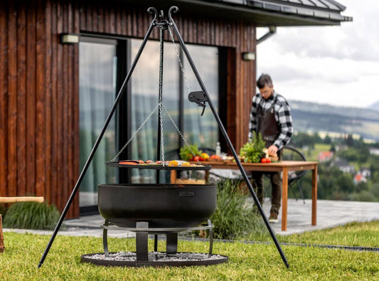 Outdoor-Grillstation mit Schwenkgrill, Viking XXL Feuerschale und runder Steinbasis auf einer Rasenfläche vor modernem Holzhaus.