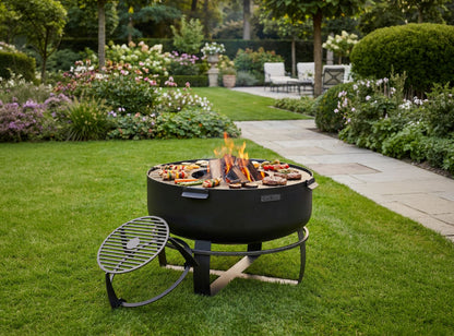 XXL Feuerschale Viking in einem gepflegten Garten mit Grillgut auf der Platte und dem Grillrost-Aufsatz im Vordergrund.