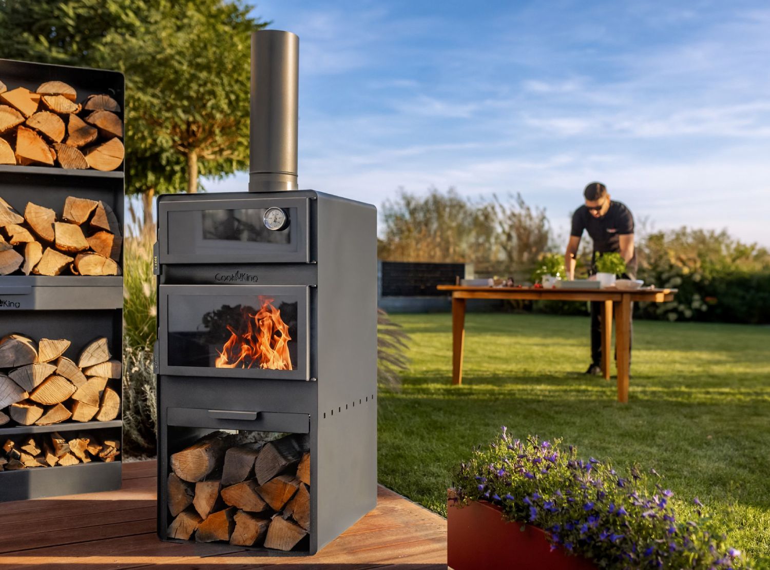Brennender Gartenofen VENTO mit Backofen und Holzregal auf einer Terrasse während der Speisenzubereitung im sonnigen Garten.
