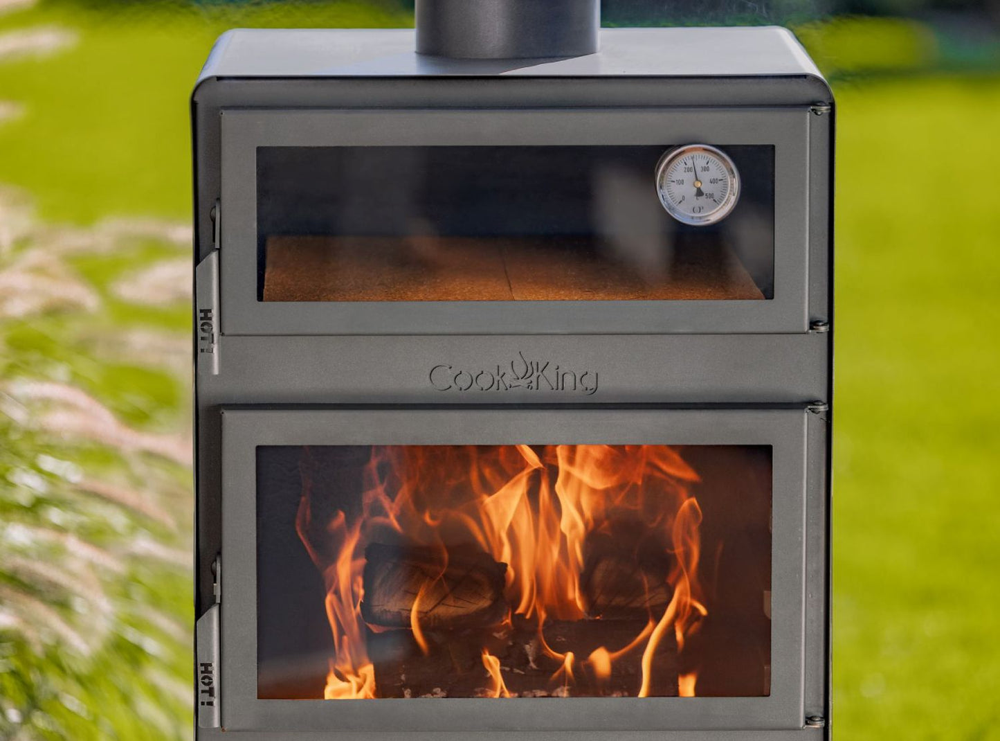 Detailansicht des brennenden Gartenofens VENTO mit sichtbaren Schamottesteinen im Backfach, integriertem Thermometer und lodernden Flammen hinter Glas.