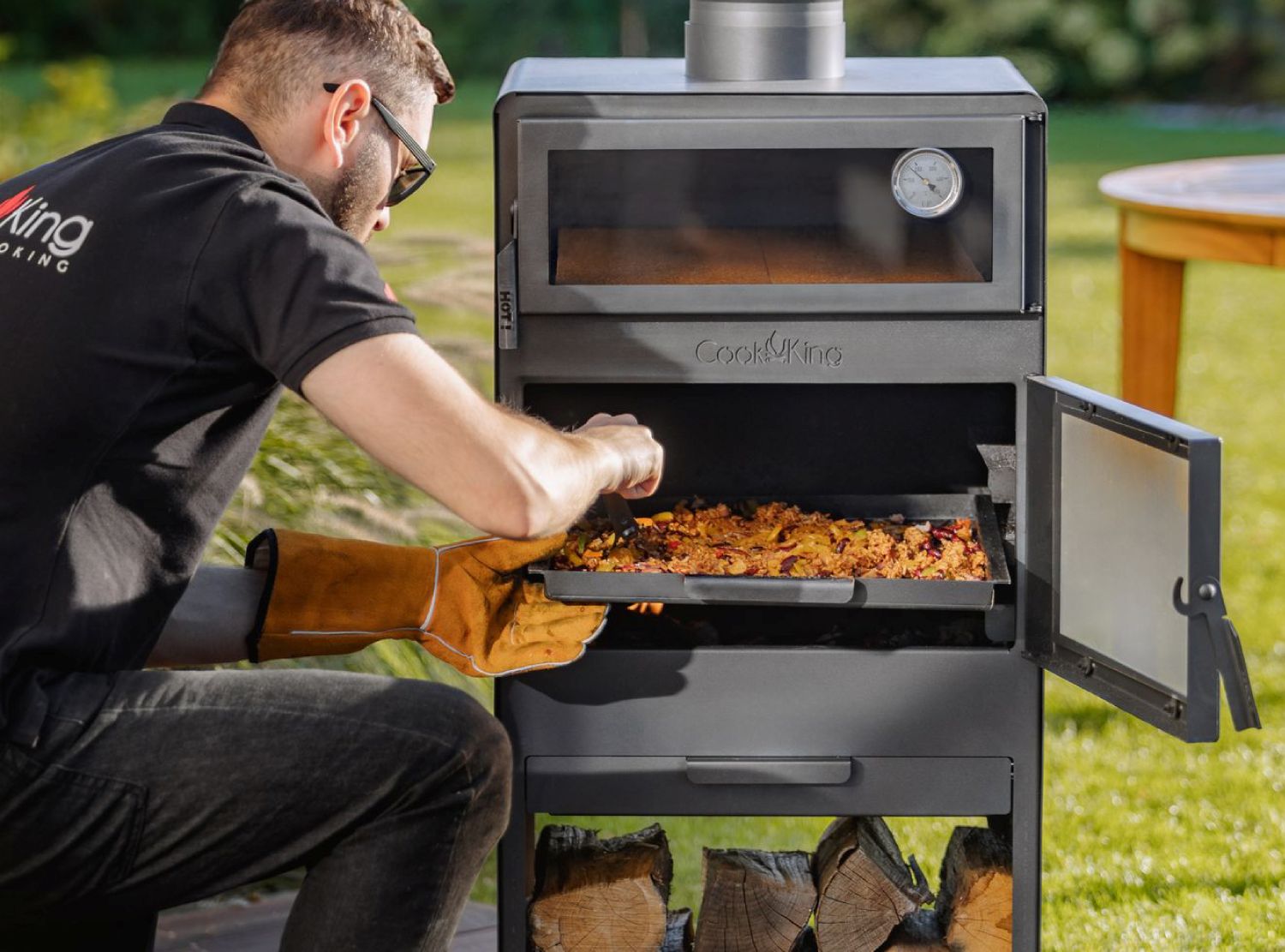 Zubereitung von Speisen im Gartenofen VENTO: Ein Mann schiebt eine Grillpfanne mit Gemüse in das offene Brennfach unter dem Backofen.