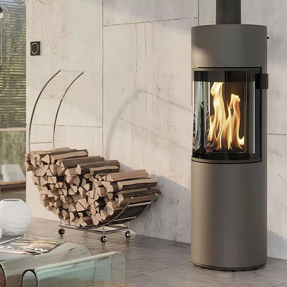 Kaminofen CERA DESIGN Prego X | 6 kW – Runder Kaminofen mit Rauchrohr neben einem Holzkorb im Wohnraum