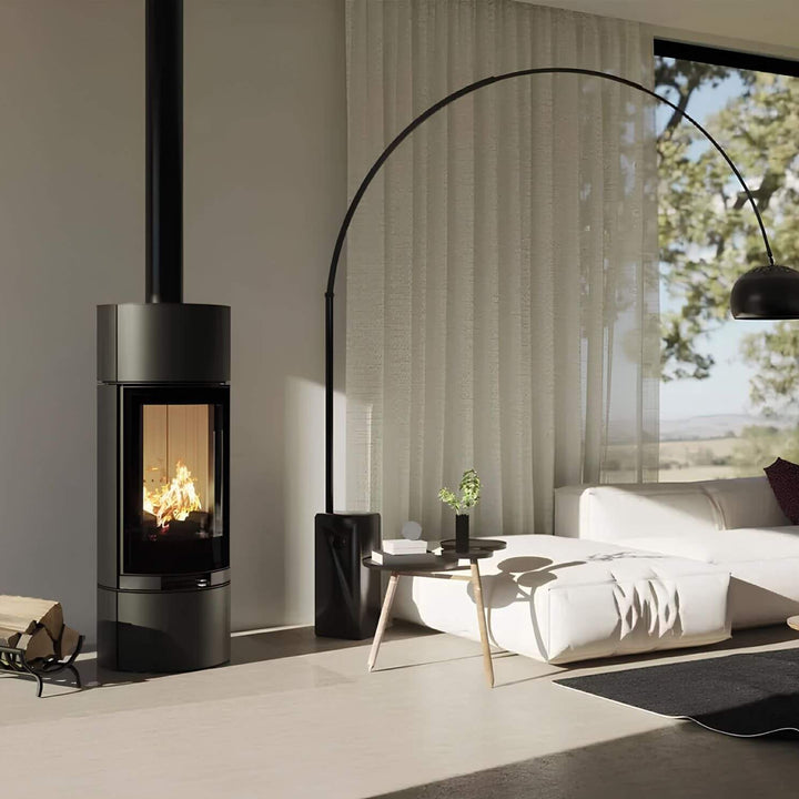 Kaminofen Hitze Charuto 8,5 kW – Hoher runder Kaminofen mit Feuer neben Sofa und Bogenlampe