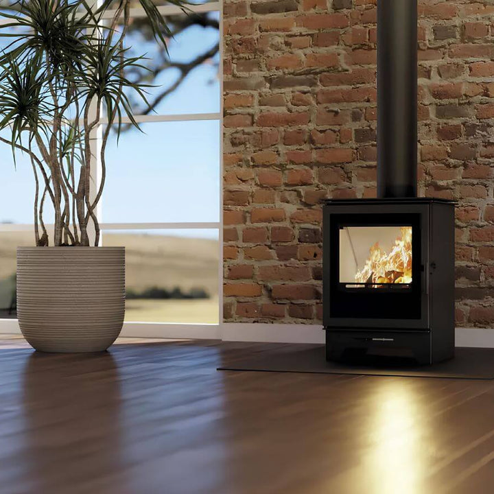 Kaminofen Hitze Gatto S 5,3 kW – Kaminofen mit Feuer vor einer Ziegelwand neben einem großen Pflanzgefäß