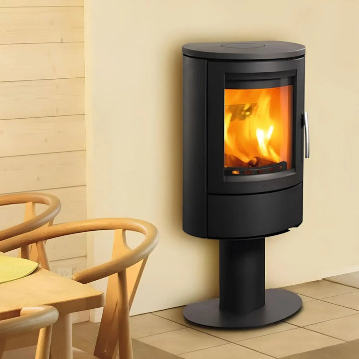 Kaminofen Varde Ovne Aura 11 | 5 kW – Kaminofen auf Standfuß mit Feuer neben einem Holzstuhl