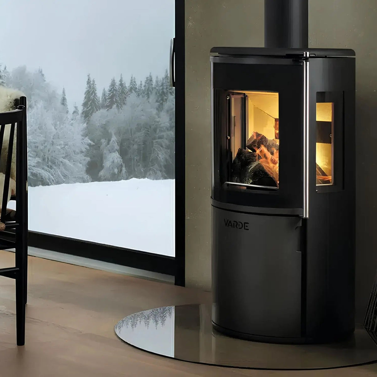 Kaminofen Varde Ovne Nordic 7 Air 6,2 kW – Kaminofen mit Feuer neben einem Fenster mit Schneelandschaft