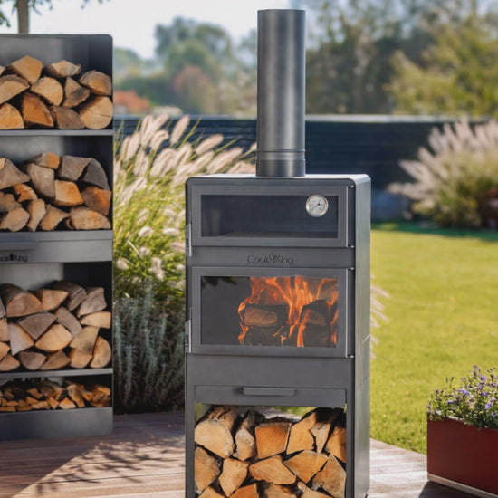 Brennender Gartenofen VENTO mit Video: Backofen und Holzregal auf einer Terrasse während der Speisenzubereitung im sonnigen Garten.