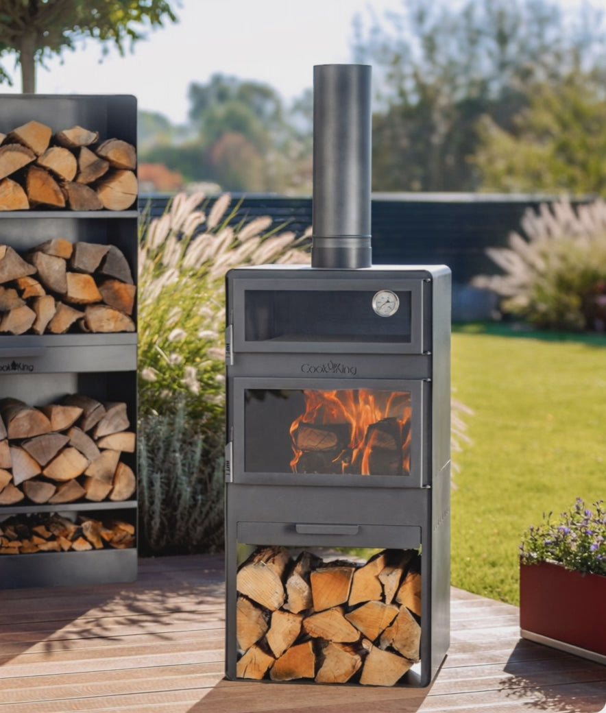 Brennender Gartenofen VENTO mit Video: Backofen und Holzregal auf einer Terrasse während der Speisenzubereitung im sonnigen Garten.